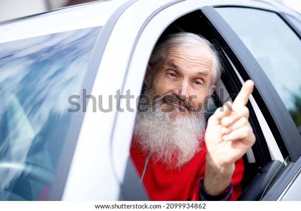 Senior man driver in car. Emotional portrait of an\
expressive and impulsive mature caucasian male driver with long\
beard, showing  his finger in air and shouting to the oncoming car\
driver