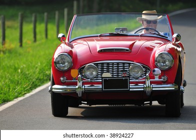 A Senior Man Driving A Classic Red Sports Car On A Country Road