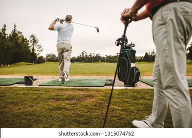 Senior man at a driving range playing golf and practice his swing with other man standing in front. Senior male golfers practicing at golf course.