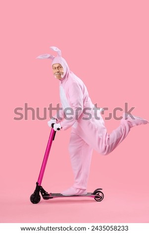 Senior man in Easter bunny costume riding kick scooter on pink background