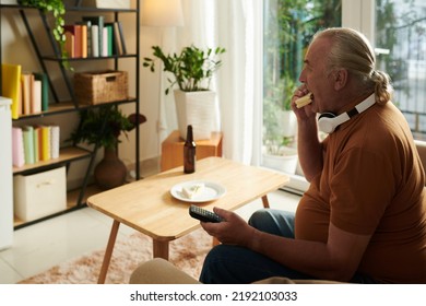 Senior Man Eating Sandwich And Watching Popular Show On Tv At Home