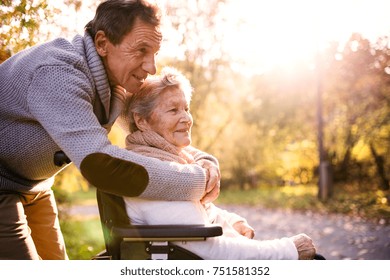 Senior Man And Elderly Woman In Wheelchair In Autumn Nature. Man With His Mother On A Walk.