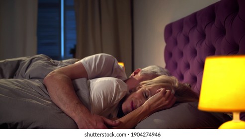 Senior man embracing beautiful asleep woman with eyes closed in bed. Side view of mature couple sleeping together in bedroom at night. Aged husband and wife sleep at home