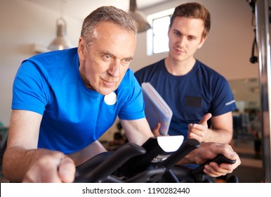 Senior Man Exercising On Cycling Machine Being Encouraged By Personal Trainer In Gym - Powered by Shutterstock