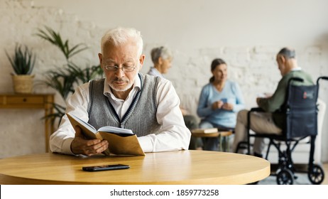 Senior man in eyeglasses reading book sitting at table in nursing home. Three elderly people, two women and disabled man, playing cards in background - Powered by Shutterstock