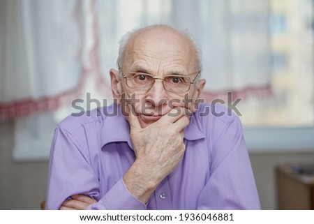Senior man in eyewear sitting at table in room against white curtain. Concept care about grandparents.