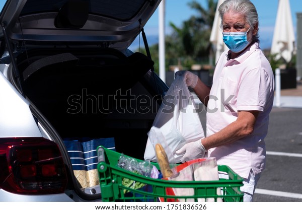 Senior man with\
face mask and protective gloves loads shopping bags from the cart\
to the trunk of the car, outdoor parking under the sun - concept of\
active retired elderly\
people