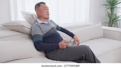 A senior man feeling bored sits on the couch, channel surfing with a remote control while unwinding alone in his bright living room during his retirement.