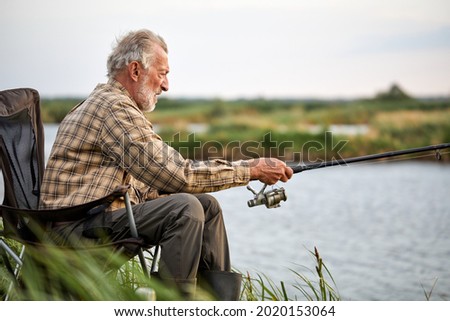 Senior man fishing outside in evening on lake in summer sitting on chair, enjoy spending time in nature, want to catch a fish, alone, side view portrait of male in casual checkered shirt
