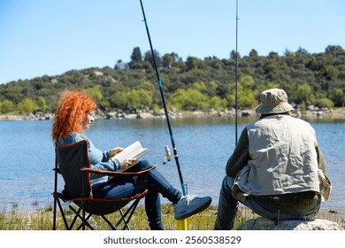 Senior man fishing and woman reading a book while sitting on a camping chair by the lake shore enjoying a sunny day - Powered by Shutterstock