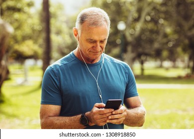 Senior man in fitness wear checking his mobile phone during workout. Portrait of a senior fitness man standing in park listening to music while working out. - Powered by Shutterstock