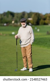 Senior Man In Flat Cap Standing With Hand In Pocket And Golf Club On Green Field