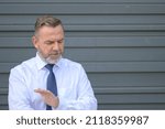 Senior man gesturing and looking down as he speaks to a person out of frame against a grey wall with copyspace