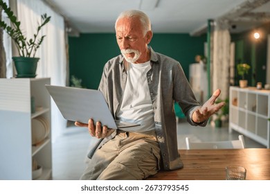 Senior man getting frustrated while working from home, holding a laptop and gesturing with his hand, showing his confusion and annoyance with technology