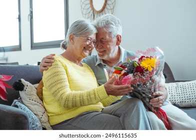 Senior man giving bouquet of flowers at his wife sitting on the sofa at home for anniversary or San Valentines’ day. Pensioners enjoying surprise together. In love people having fun. - Powered by Shutterstock