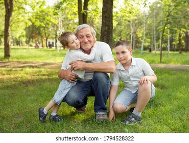 Senior Man And Grandsons Are Hugging And Smiling While Resting Together In The Garden. Men Day. Joyful Grandfather Spending Time With His Grandkids. Man Playing With Grandchildren. Real Life, Family