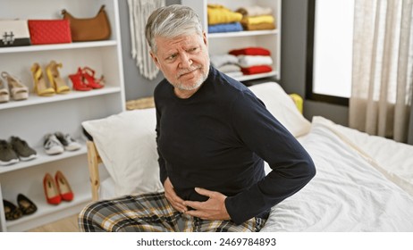 Senior man with gray hair experiencing stomach pain while sitting on a bed in a well-lit bedroom - Powered by Shutterstock