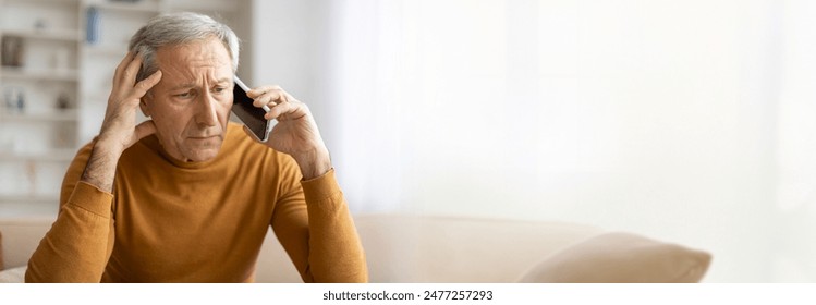 A senior man with gray hair sits on a couch in his living room, talking on the phone with a worried expression on his face. He appears concerned and is holding his hand to his forehead, copy space - Powered by Shutterstock