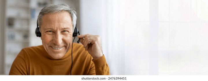 A senior man with gray hair is wearing a headset and smiling at the camera. He is likely working from home, as he is sitting in front of a window, copy space - Powered by Shutterstock