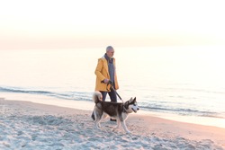 Senior Man With Grey Hair And Beard In A Yellow Raincoat With Husky Dog On Leash Walking Along Sea On The Beach At Sunrise
