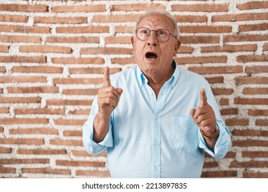 Senior Man With Grey Hair Standing Over Bricks Wall Amazed And Surprised Looking Up And Pointing With Fingers And Raised Arms. 