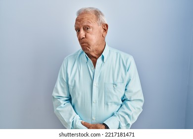 Senior man with grey hair standing over blue background puffing cheeks with funny face. mouth inflated with air, crazy expression. 