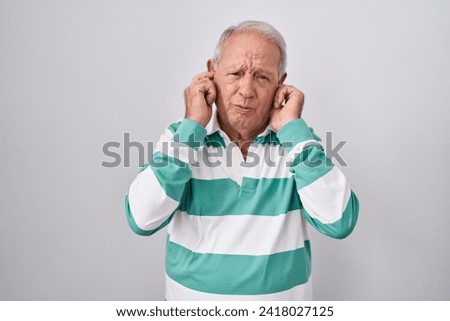 Senior man with grey hair standing over white background covering ears with fingers with annoyed expression for the noise of loud music. deaf concept. 