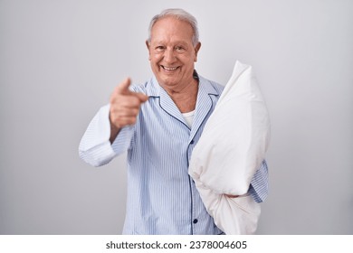 Senior man with grey hair wearing pijama hugging pillow pointing fingers to camera with happy and funny face. good energy and vibes. 