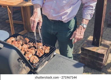 A Senior Man Is Grilling Pieces Of Chicken On A Barbecue In A Garden On A Sunny Day