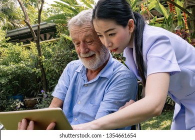 Senior Man Happiness Sitting On Wheelchair With Smiling Nurse, Takes Care And Discussion And Cheer In The Garden At Nursing Home, Using Tablet Or Mobile For Selfies Or Learning Technology