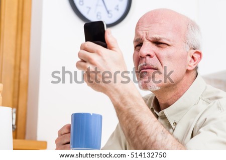 senior man has problems with his vision while trying to read or watch something on the screen of his mobile phone in his kitchen