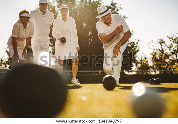 Senior man in\
hat bending forward to throw a boules in a lawn. Elderly man\
playing boules in a lawn with his\
friends.