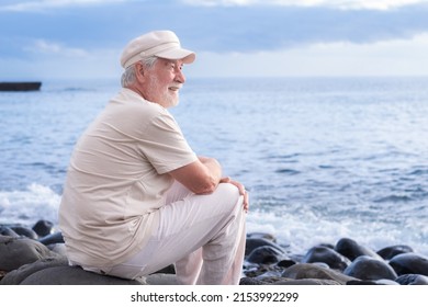 Senior Man With Hat Sitting On The Beach Relaxing Enjoying Retirement. Elderly Bearded Male Enjoying Sunset At Sea, Horizon Over Water