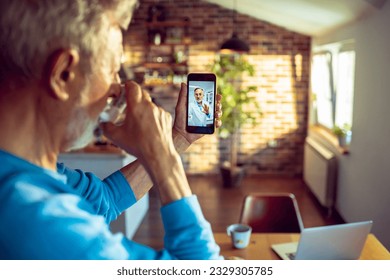 Senior man having a video call with his doctor on the smart phone in the kitchen while having breakfast - Powered by Shutterstock