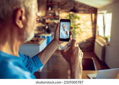 Senior man having a video call with his doctor on the smart phone in the kitchen while having breakfast - Powered by Shutterstock