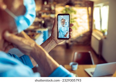 Senior man having a video call with his doctor on the smart phone in the kitchen while having breakfast - Powered by Shutterstock