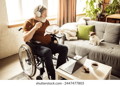 Senior man in headphones sitting on wheelchair at home in living room with dog, watching movie on laptop and embroidery. Concept of healthcare, lifestyle, wellness, comfort, empowerment - Powered by Shutterstock