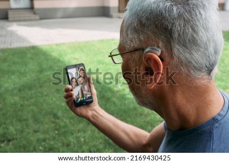 Senior man with a hearing aid behind the ear communicates with his family via video communication via a smartphone. Full human life with hearing aids