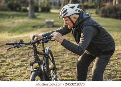 A senior man with a helmet is intently inspecting his mountain bike handlebars in a park setting. - Powered by Shutterstock