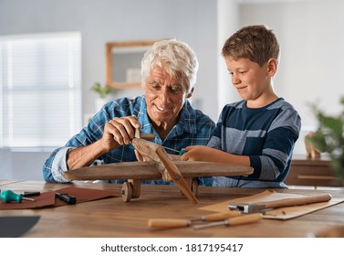 Senior Man Helping Child To Screw An Airplane Part That They Are Building Together During Summer Vacation. Retired Grandfather Helping Grandson In Making Wooden Plane At Home For School Project.