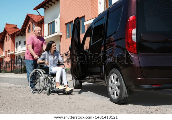 Senior man helping young woman in wheelchair to\
get into van outdoors