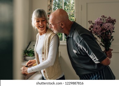 Senior man hiding a bunch of flowers behind him to surprise his wife. Woman cutting vegetables in kitchen while her husband standing behind her with a bunch of flowers. - Powered by Shutterstock