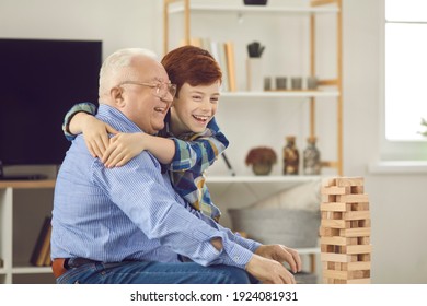 Senior Man With His Grandchild Having Fun At Home. Happy Smiling Grandson Embracing His Grandfather While Playing Together. Little Kid Laughing And Hugging Grandad While Playing Tumble Tower