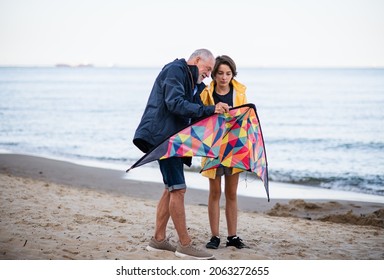 Senior man and his preteen granddaughter preparing kite for flying on sandy beach.