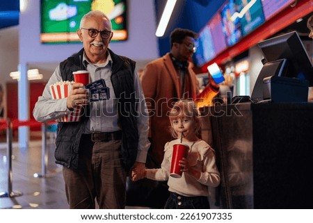 A senior man with his small granddaughter is in the movie hall going to see a movie and having popcorn and drinks.