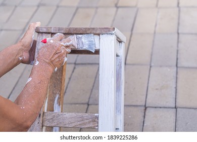 Senior man holding brush for paint white on old wooden chair without a backrest Hand of man. Old chairs are painted white. Renovation of old furniture