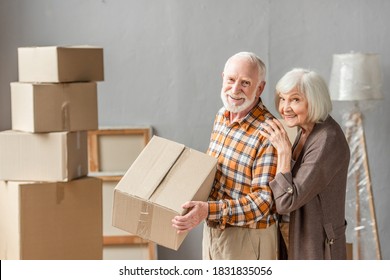 Senior Man Holding Cardboard Box And Wife Leaning On Shoulder In New House, Moving Concept