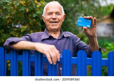 Senior Man Holding Credit Card In Hand And Looking In Camera While Standing Beside His Country House.