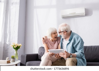 Senior man holding digital tablet and embracing smiling wife on couch