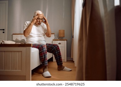 Senior man holding his head in pain while sitting on a bed at home. Copy space. - Powered by Shutterstock